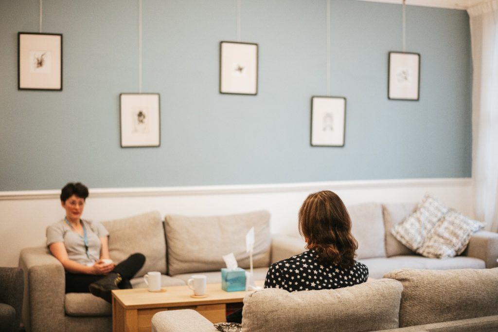 Interior, two women in a living room chatting over cups of tea, for “Kyra’s Drop-In Sessions: A Place to Simply Be”