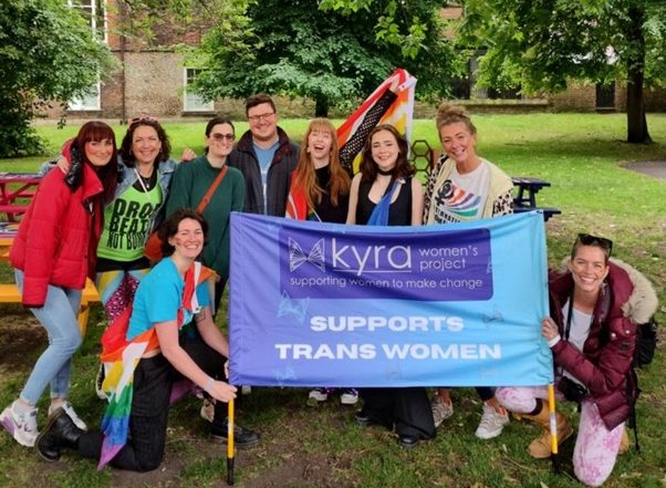 Exterior, a group of women wearing rainbow flags and holding a sign reading Kyra Supports Trans Women, for “A New LGBTQ+ Women’s Group at Kyra”
