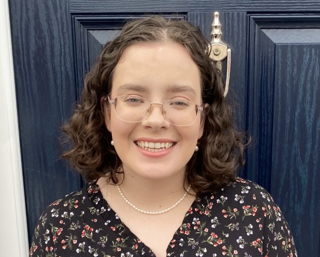 Exterior, a young smiling woman stood in front of a door, for “Christina’s Journey: Why ROSE Matters”