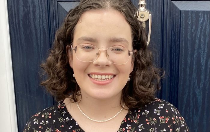 Exterior, a young smiling woman stood in front of a door, for “Christina’s Journey: Why ROSE Matters”