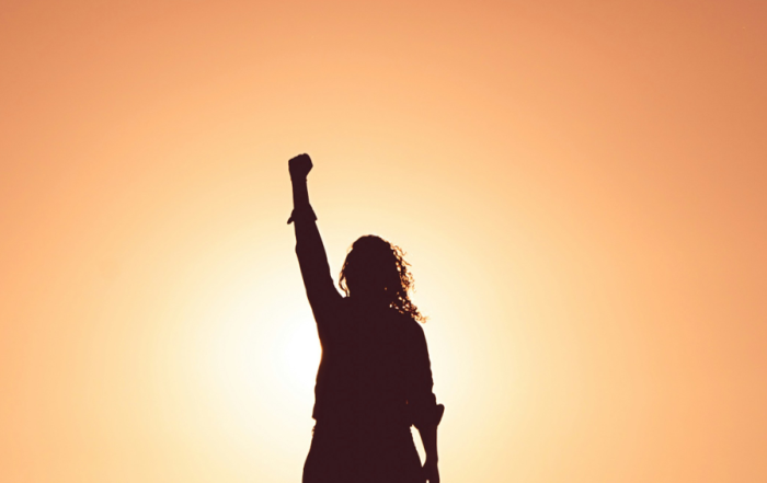Exterior, a woman holding her fist aloft against a magnificent orange sunset, for “Meet Ashley: From Member to Trustee”