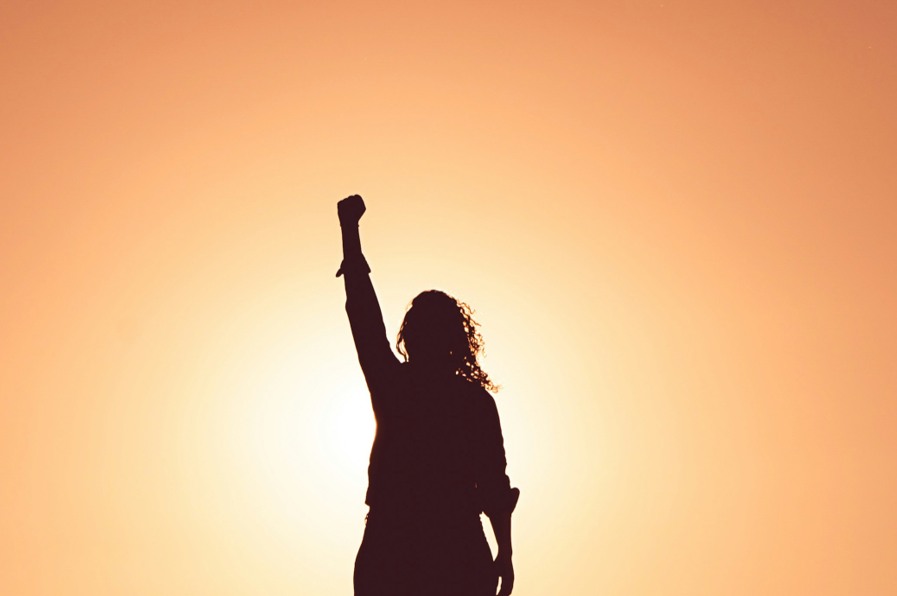 Exterior, a woman holding her fist aloft against a magnificent orange sunset, for “Meet Ashley: From Member to Trustee”
