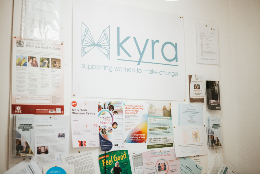 Interior, a corkboard reading “Kyra” with many different leaflets and posters attached, for “From Surviving to Reclaiming: A FREYA Journey”